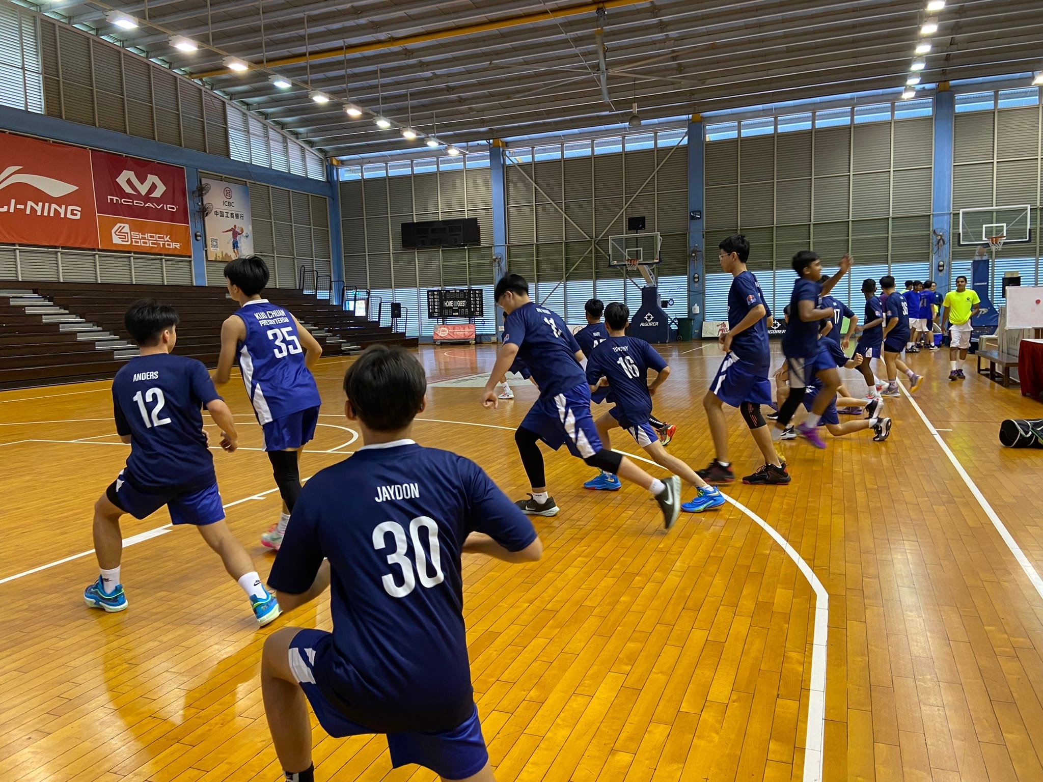 Young basketball players in blue numbered jerseys warm up on a wooden indoor court with sponsor banners on the wall.